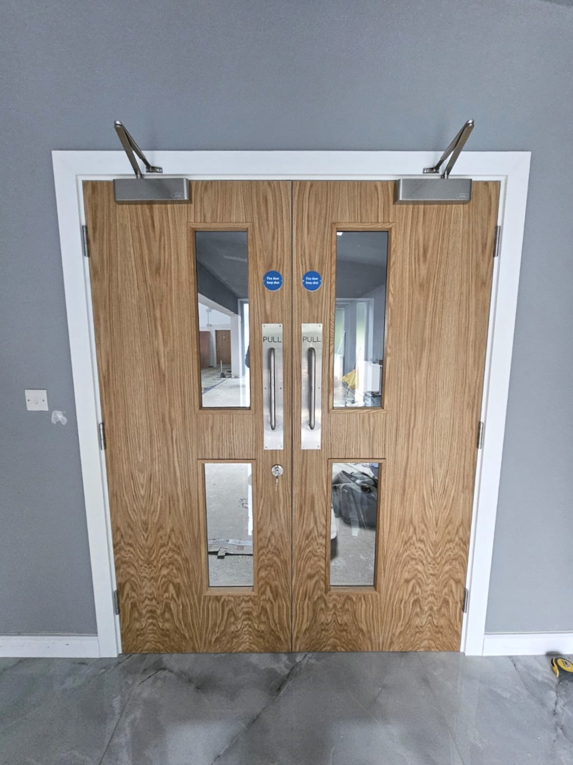 Brown timber commercial double fire doors with glazed vision panels, pull handles and overhead closers