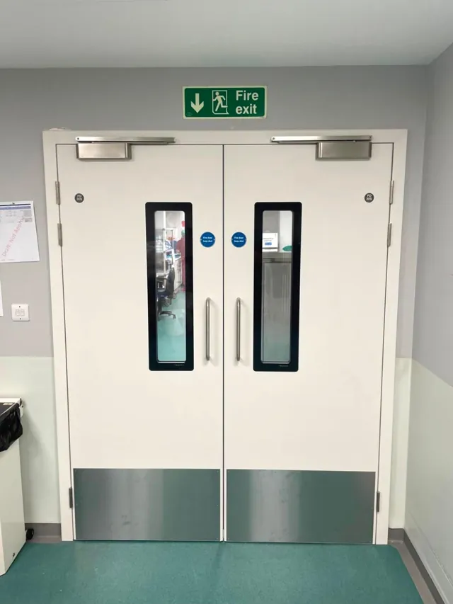 White double doors with vision panels and green lower panels viewed from the corridor
