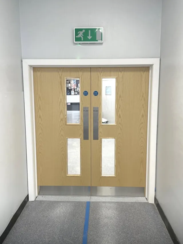 Pale timber double doors with vertical glazing below a green exit sign