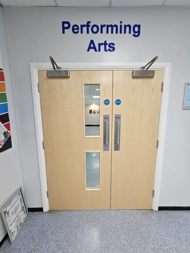Pale timber double doors with vision panels beneath a Performing Arts sign